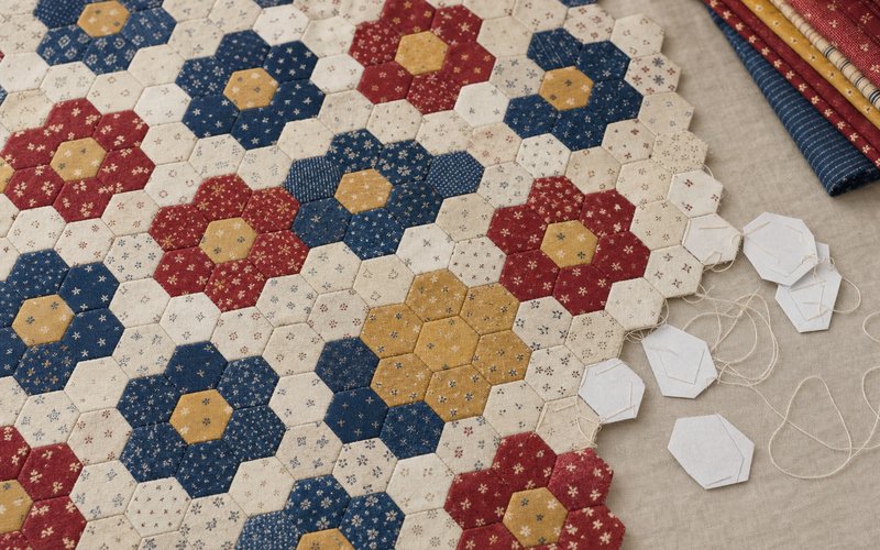 English paper-pieced hexagons arranged as a grandmother’s flower garden in indigo, madder and ochre, with paper templates and basting thread at the edge.