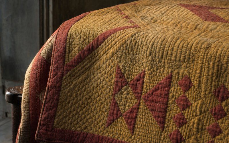 An antique Welsh wool quilt in ochre and faded red, its surface covered in dense hand-stitched geometric quilting.